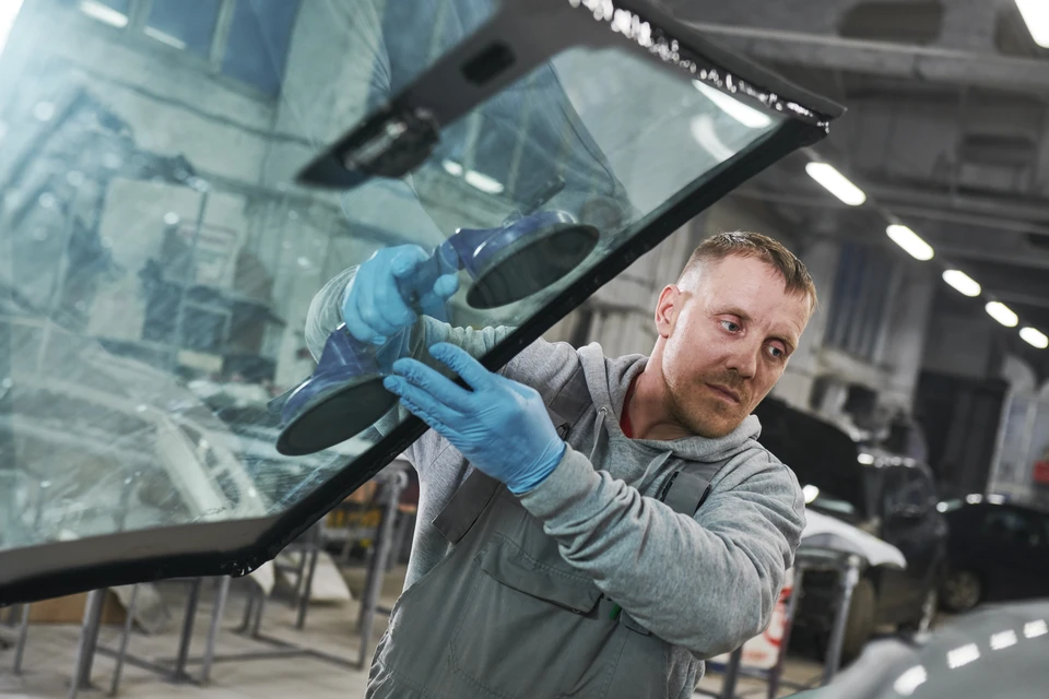 Atelier de réparation automobile intérieur avec technicien en combinaison grise et gants de protection bleus installant un pare-brise sur véhicule