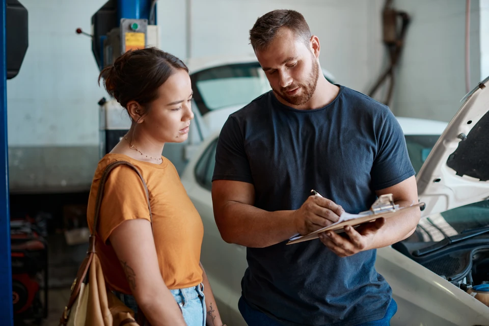 Photo professionnelle en intérieur d'atelier automobile : un véhicule utilitaire ou une berline professionnel en cours d'intervention pour réparation de vitrage, avec un technicien en tenue de travail visible effectuant une réparation ou un remplacement de pare-brise. L'environnement d'atelier moderne et bien équipé (établi, outils de réparation, système d'éclairage professionnel) doit être visible en arrière-plan pour contextualiser le cadre B2B et la prise en charge professionnelle des flottes de véhicules.