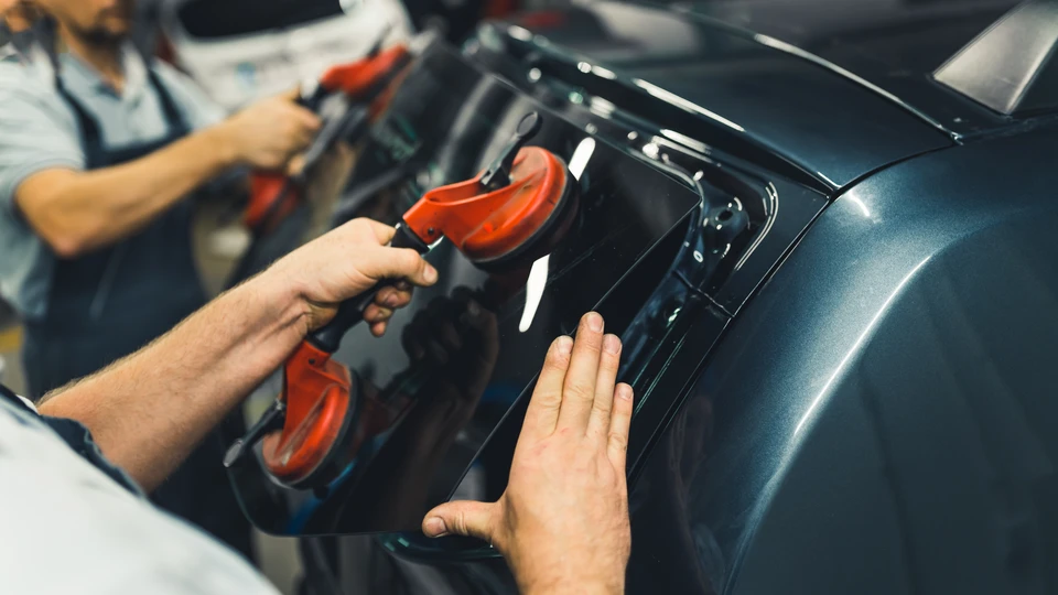 Photographie professionnelle montrant un client remettant les clés de son véhicule à un technicien Vitrauto 14 dans un atelier de réparation automobile. Le client sourit avec satisfaction, le technicien porte l'uniforme/gilet de l'entreprise et tient un document (devis ou prise en charge d'assurance). Le véhicule endommagé est visible en arrière-plan dans l'atelier. Environnement professionnel avec éclairage de garage, établis et équipements visibles. L'image transmet la confiance, la prise en charge sans souci et la relation client satisfaite.