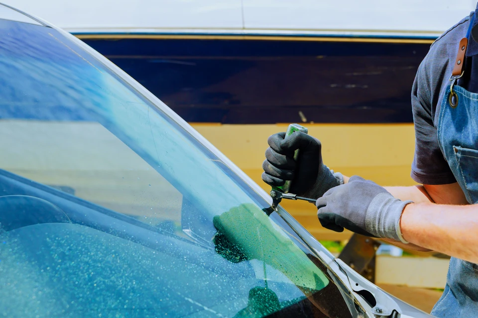 Technicien en combinaison bleue effectuant le remplacement d'un pare-brise automobile à l'extérieur, utilisant des ventouses de manutention professionnelles pour manipuler le vitrage