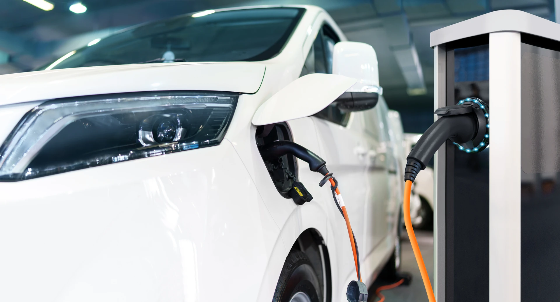 Photographie professionnelle d'un véhicule électrique ou hybride de marque reconnue (Tesla Model 3, Peugeot e-3008, Renault Zoe, ou équivalent) en cours de remplacement de pare-brise dans l'atelier de Vitrauto 14. Le véhicule est positionné face avant visible, pare-brise en cours de dépose ou fraîchement remplacé. On aperçoit un technicien en combinaison bleue effectuant les finitions ou les joints de calfeutrage. L'arrière-plan montre l'atelier avec son équipement caractéristique (établis, outils de pose, éclairage professionnel). La scène illustre clairement la spécialité « véhicules électriques et hybrides » avec soin apporté aux systèmes connectés et aux particularités de ces véhicules modernes.