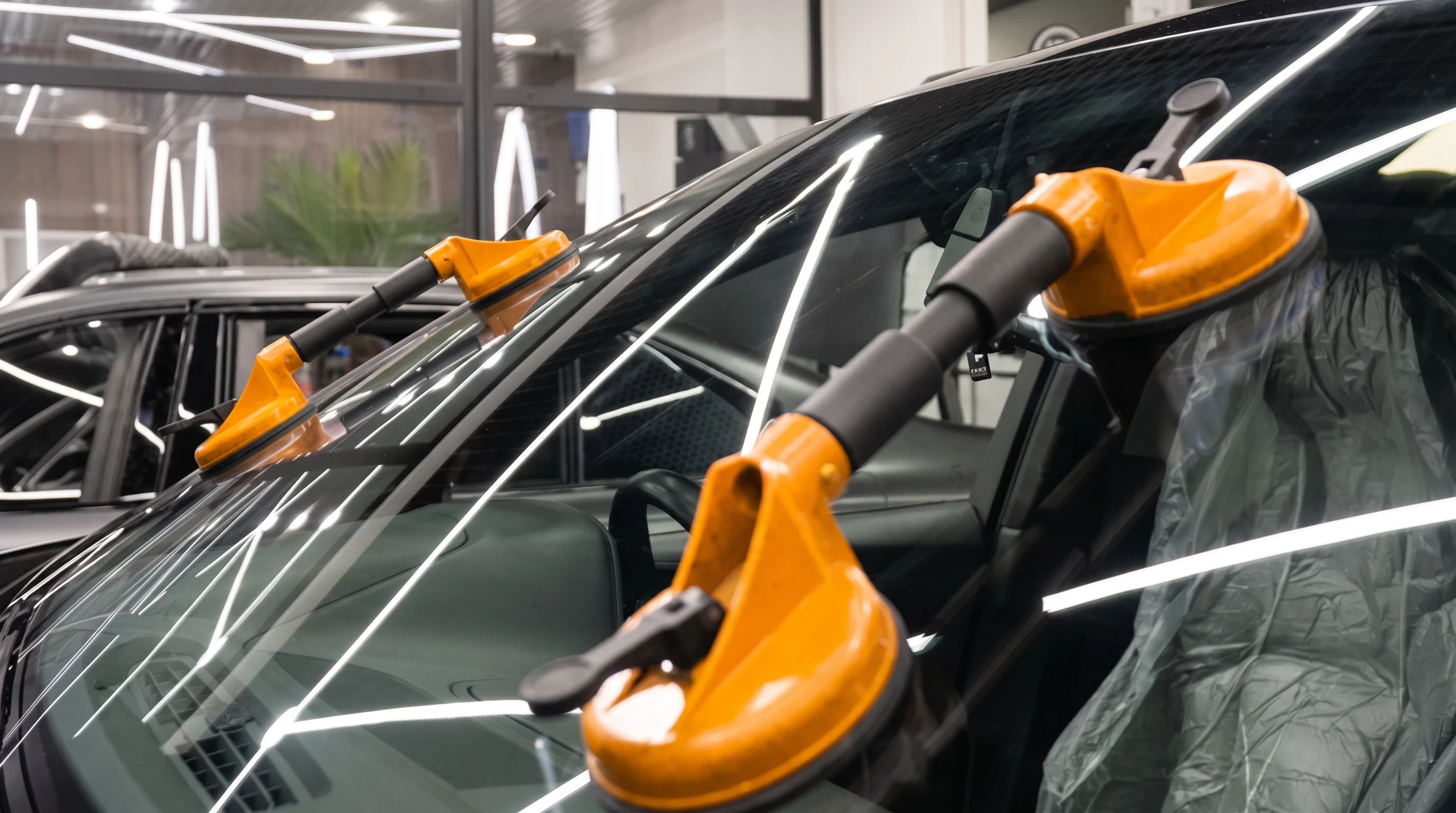 Photo réaliste professionnelle dans un atelier de vitrage automobile propre et lumineux, montrant un technicien en tenue de travail sobre en train de réparer un impact sur le pare-brise d’une voiture à l’aide d’un injecteur de résine et d’une lampe UV. Le véhicule est partiellement visible de face, le capot protégé par une housse, avec des outils spécialisés disposés proprement à proximité. Ambiance sérieuse, moderne et rassurante, cadrage large adapté à une bannière de site web.